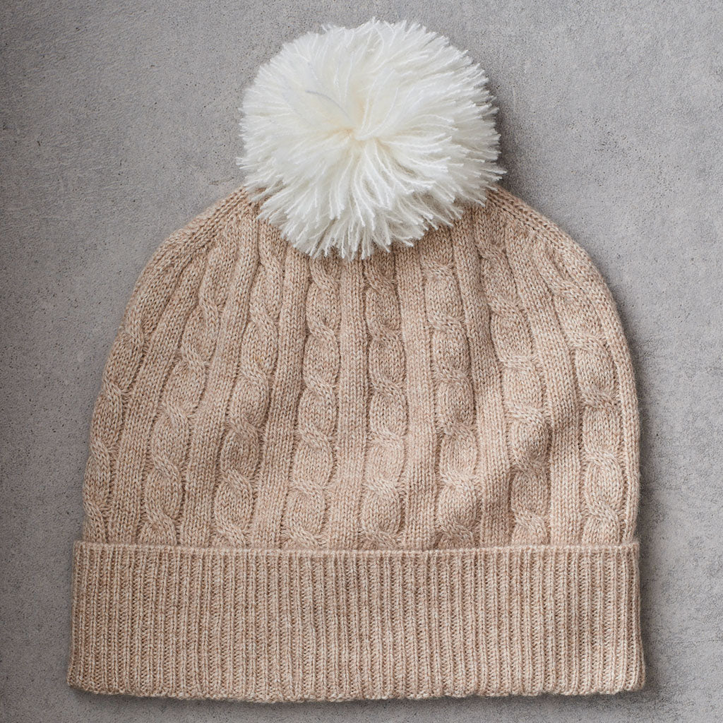 A beige cable-knit beanie hat with a wide ribbed brim and a fluffy white pom-pom on top, laid flat on a textured gray background.