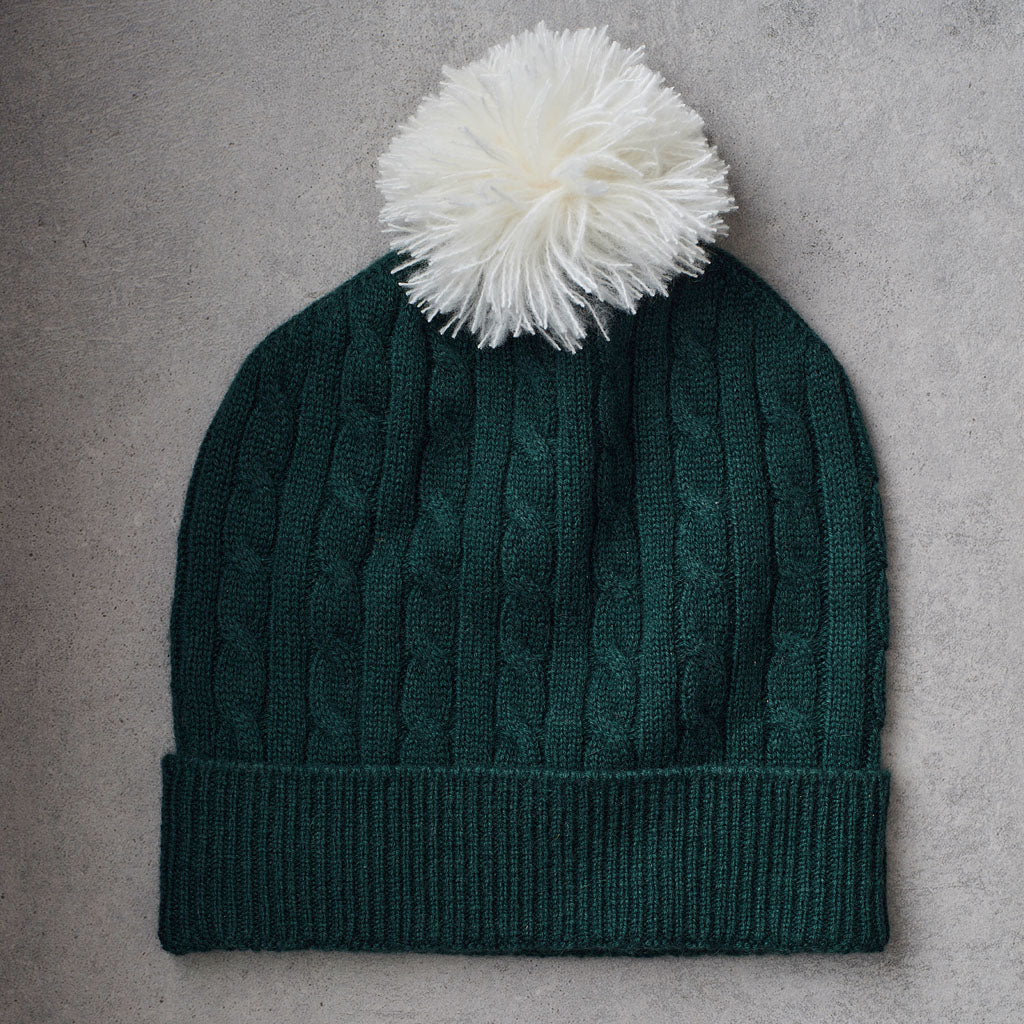 A dark green cable-knit beanie with a ribbed cuff and a fluffy white pom-pom on top, laid flat on a textured gray background.