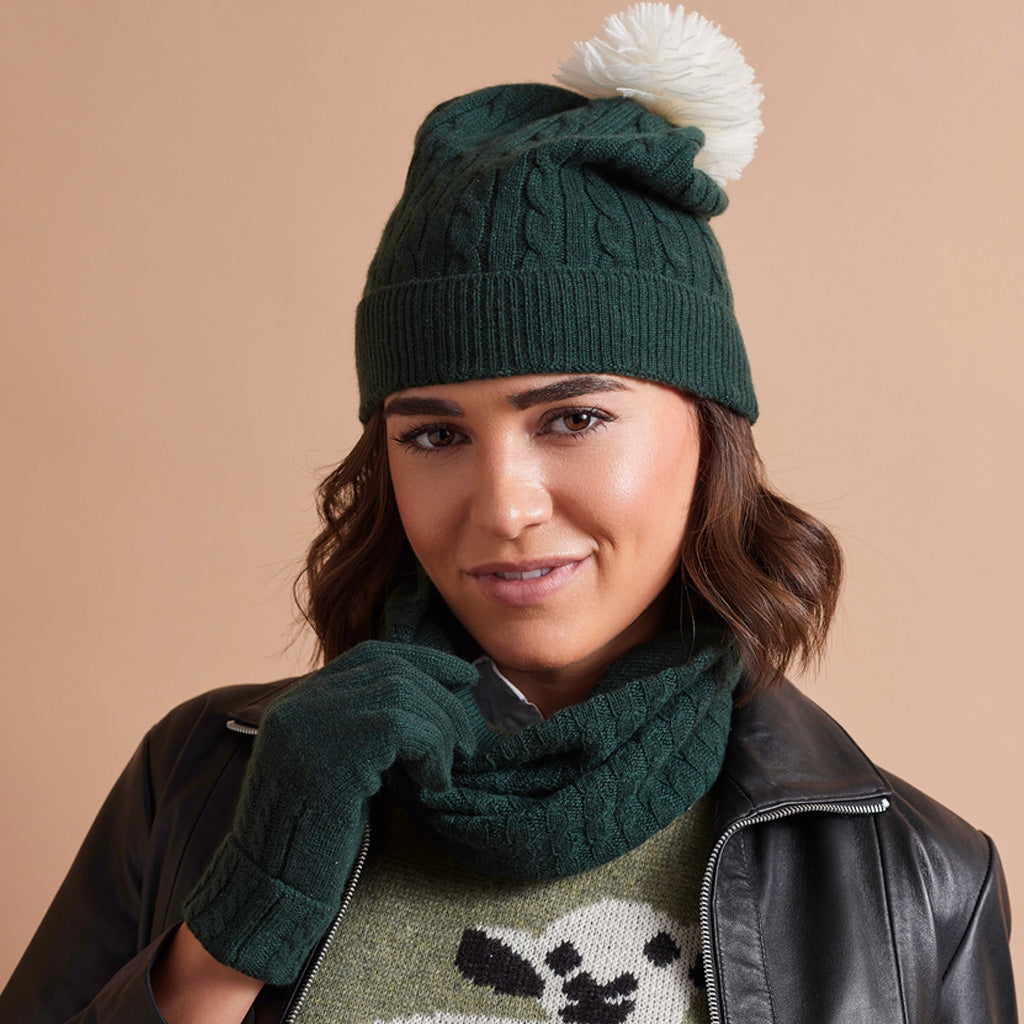 A woman wearing a dark green cable-knit beanie with a fluffy white pom-pom, a matching dark green knit scarf, and a black leather jacket against a solid beige background.