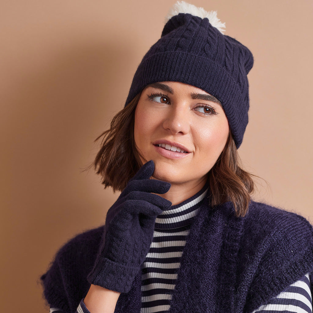 A woman wearing a navy blue cable-knit beanie with a white pom-pom, matching navy knit gloves, and a navy knit sweater vest over a black and white striped turtleneck, posed against a solid warm beige background.