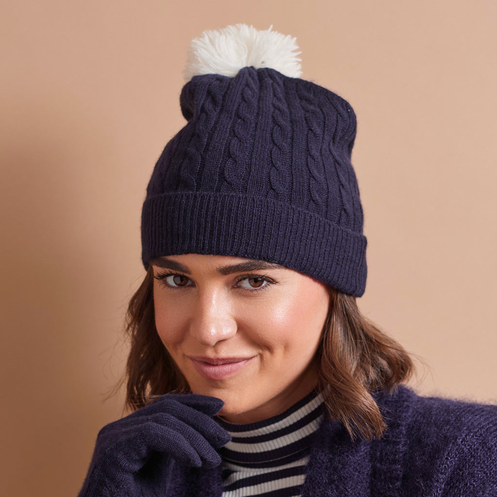 A woman wearing a navy blue cable-knit beanie with a white pom-pom, matching navy knit gloves, and a navy knit sweater vest over a black and white striped turtleneck, posed against a solid warm beige background.