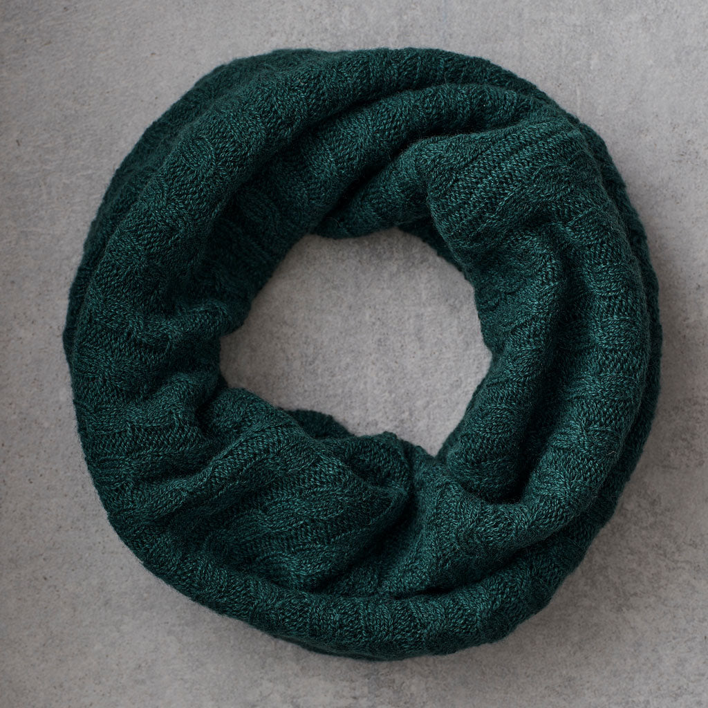 A dark green ribbed knit infinity scarf, coiled into a circle, displayed on a neutral gray textured background.