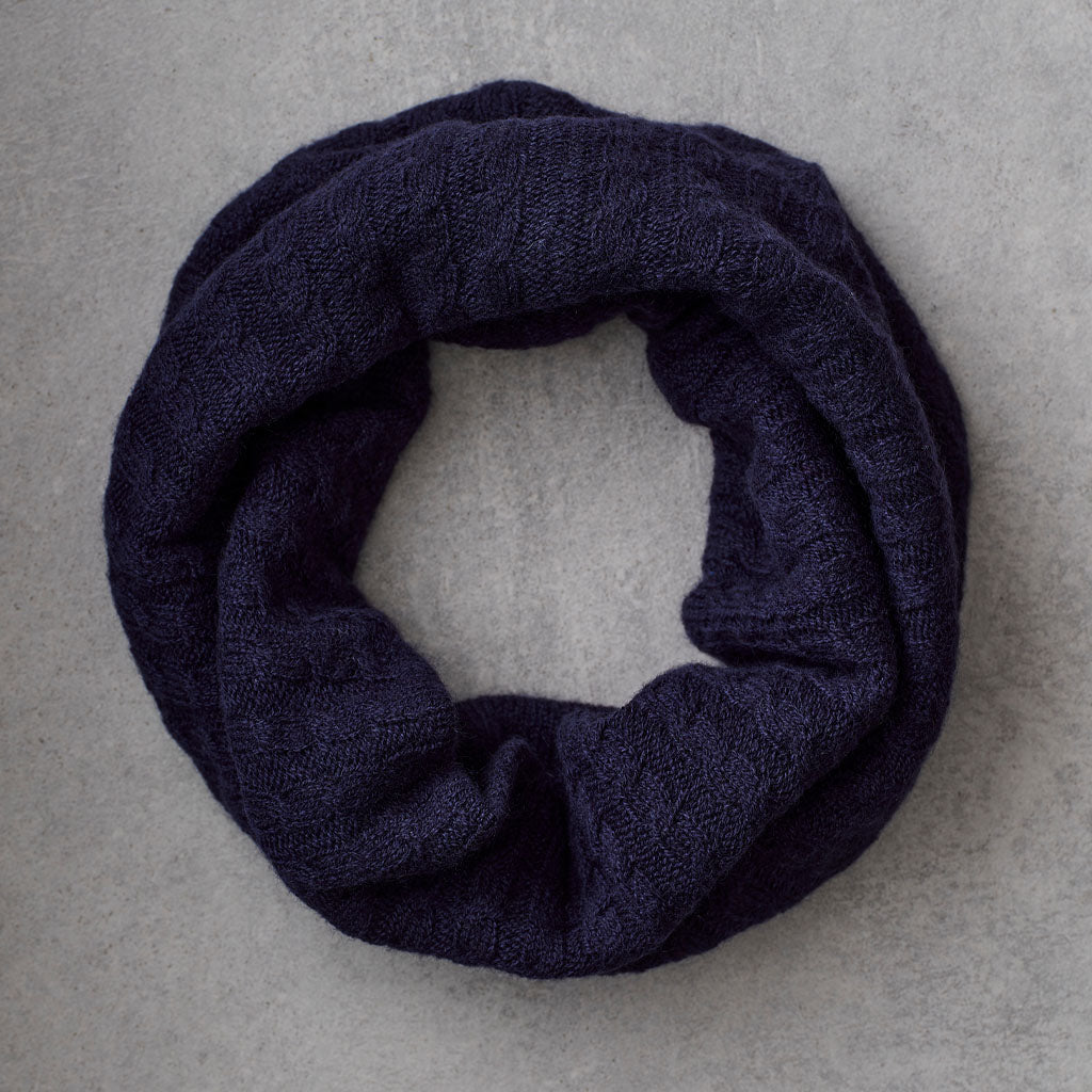 A dark navy blue textured neckwarmer, folded into a ring, displayed on a neutral gray background.