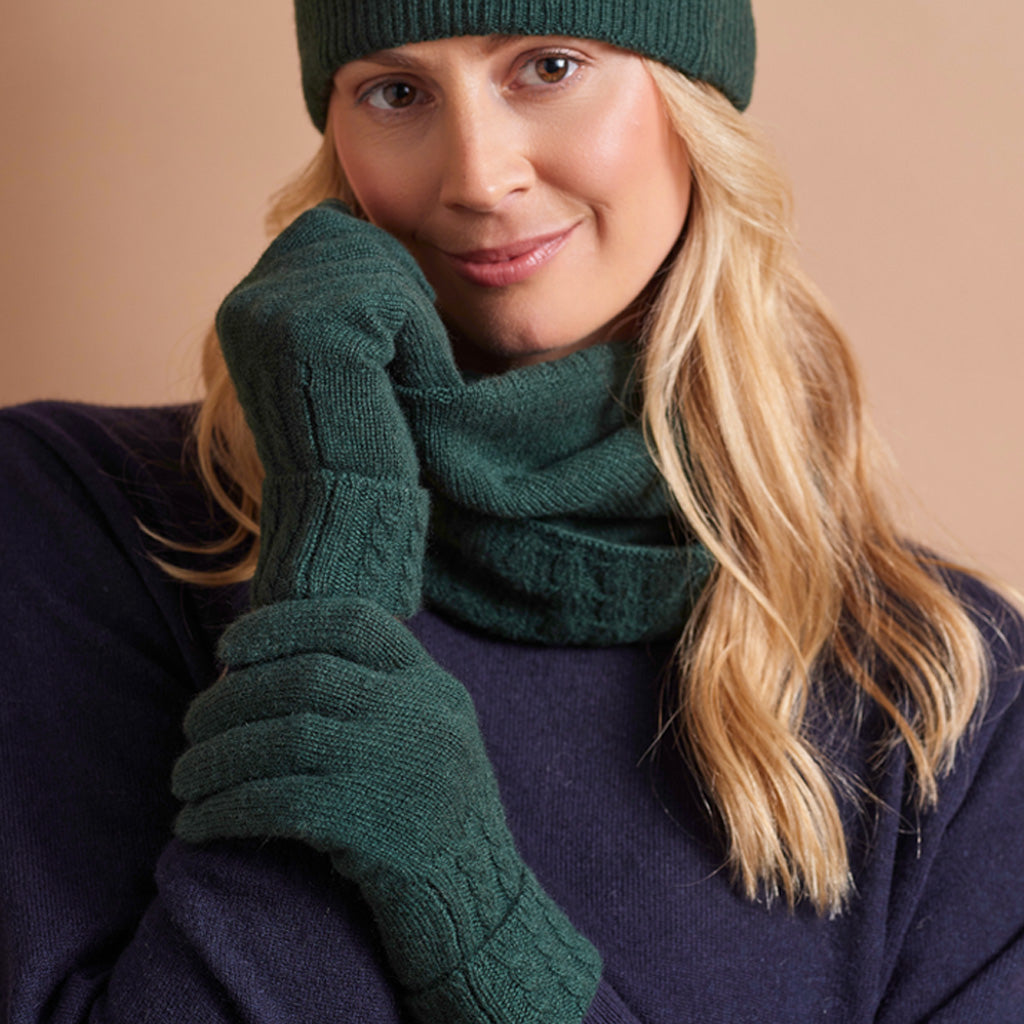 A woman wearing a dark green knit beanie, matching knit gloves, and a matching knit neck warmer, paired with a dark navy sweater against a solid beige background.