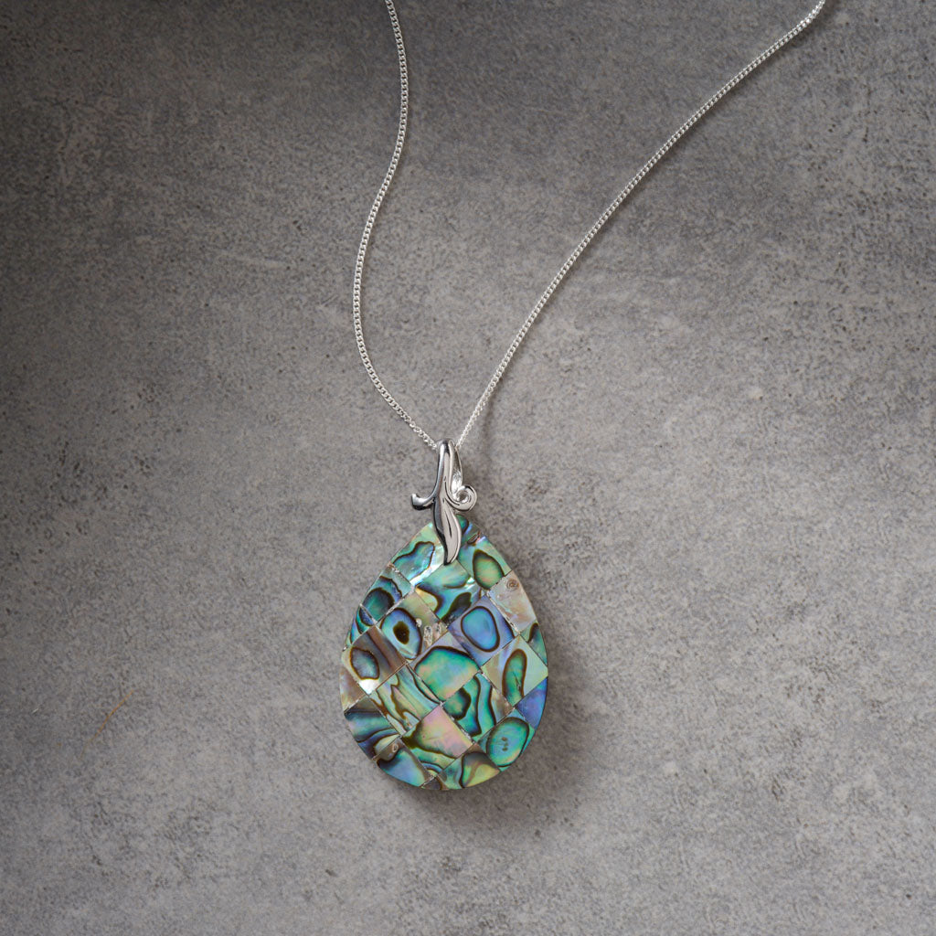 A silver chain necklace with a teardrop-shaped pendant made of iridescent abalone shell, displayed on a textured gray background.
