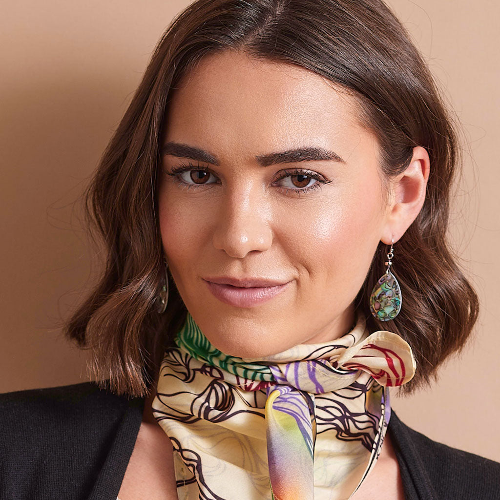 A woman with wavy brown hair wearing a beige silk scarf with a colorful abstract pattern and silver teardrop earrings, set against a solid beige background.