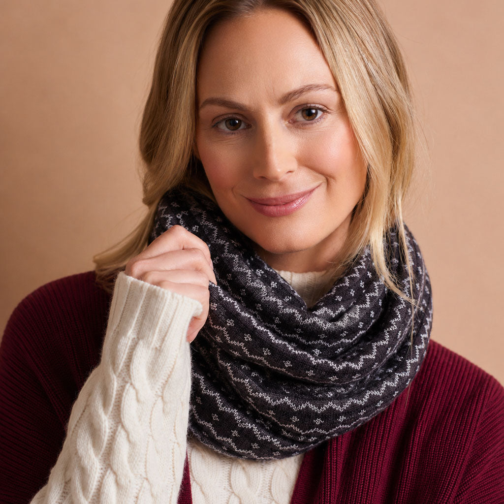 A woman wearing a dark charcoal gray infinity scarf with a white geometric pattern, styled with a burgundy sweater and a cream cable-knit top against a solid beige background.