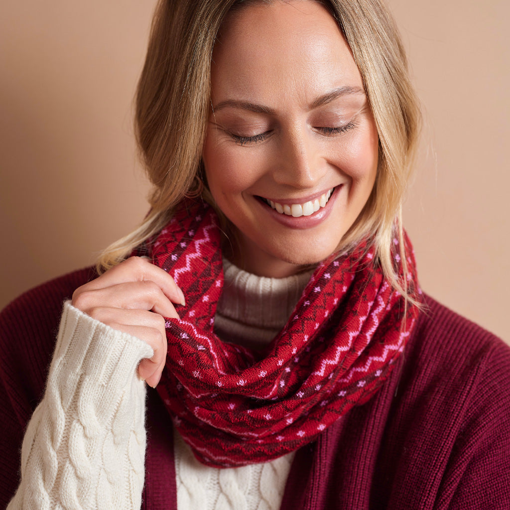 A woman wearing a red infinity scarf with a white geometric pattern, styled with a burgundy sweater and a cream cable-knit top against a solid beige background.