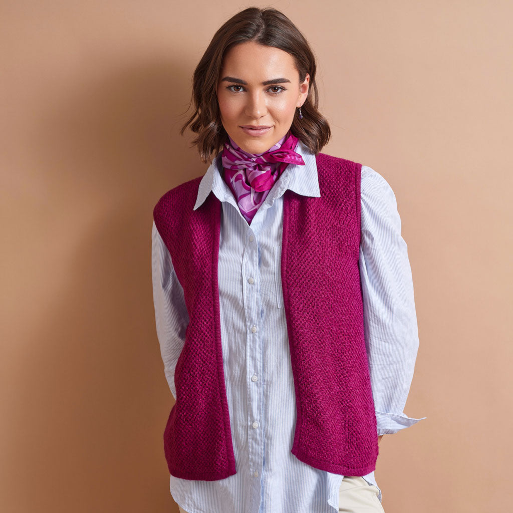 A woman wearing a sleeveless, textured magenta knit vest over a light blue button-down shirt, accessorized with a pink patterned neck scarf, set against a solid warm beige background.