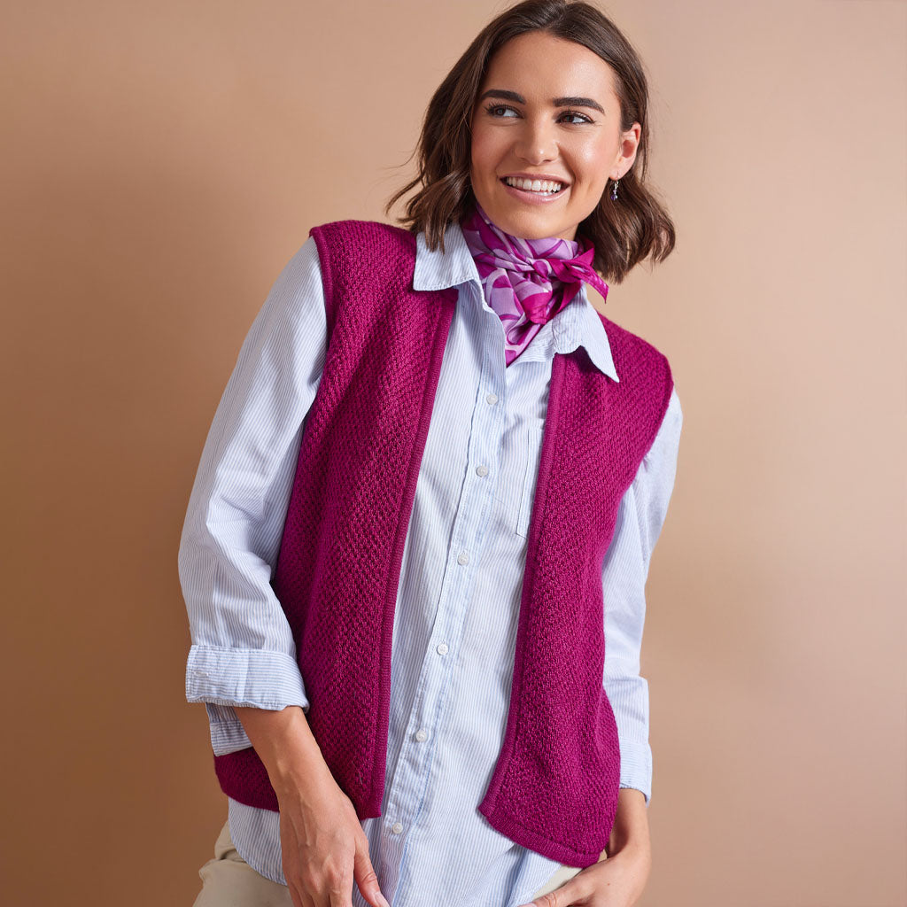 A woman wearing a bright magenta knit vest over a light blue striped button-down shirt, accessorized with a pink patterned neck scarf, posed against a solid warm beige background.