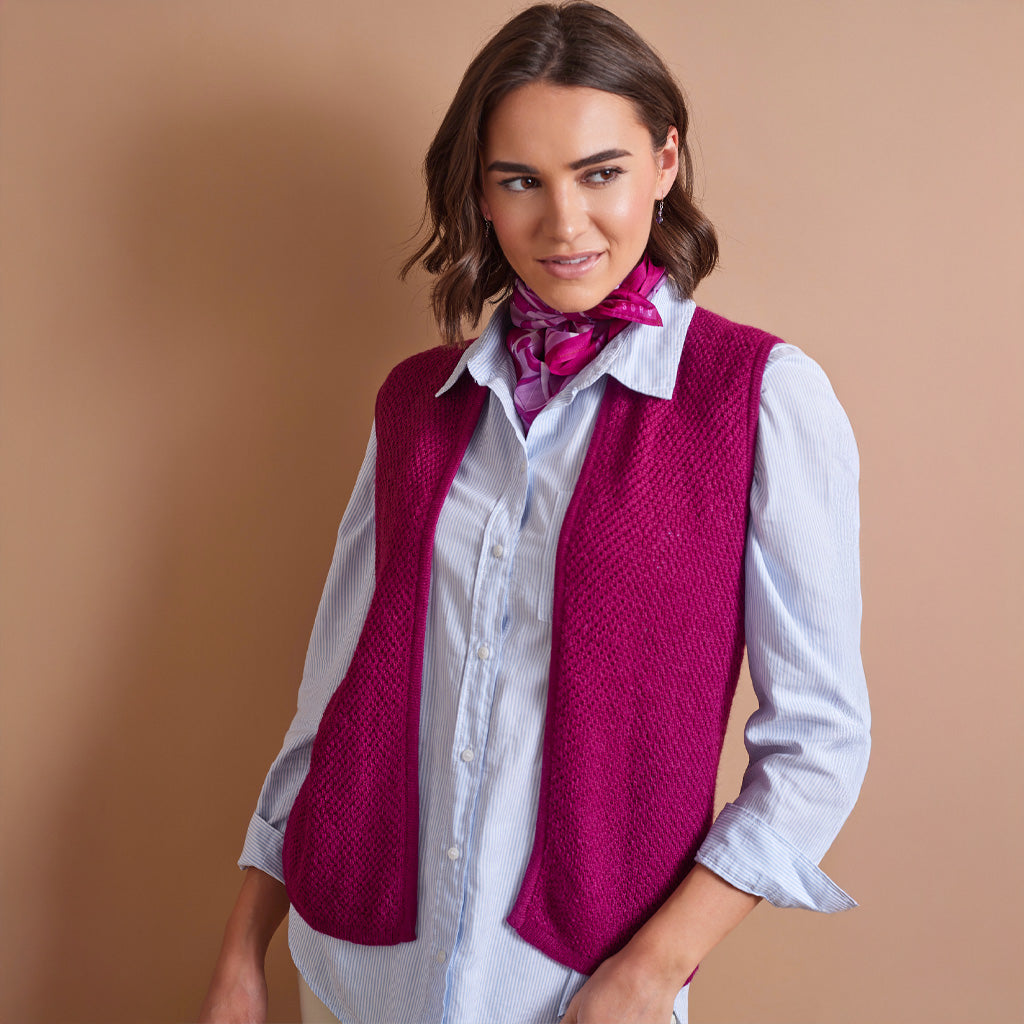 A woman wearing a bright magenta knit vest over a light blue striped button-up shirt, accessorized with a pink patterned neck scarf, posed against a solid warm beige background.