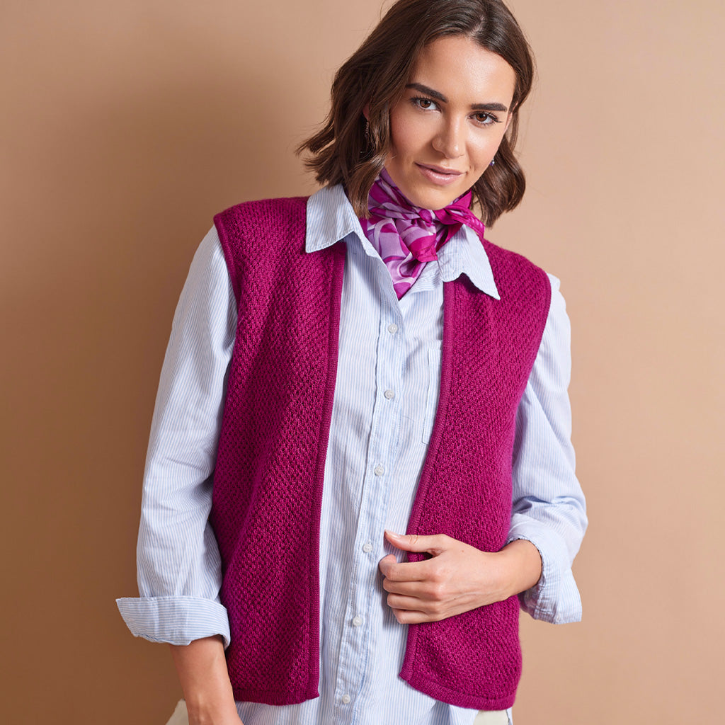 A woman wearing a bright magenta knit sleeveless vest over a light blue striped button-up shirt, accessorized with a matching pink patterned neck scarf, set against a solid warm tan studio background.