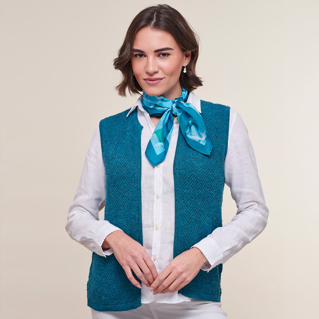A woman wearing a teal blue textured knit vest over a white button-up shirt, accessorized with a bright blue patterned silk neck scarf, against a solid beige background.
