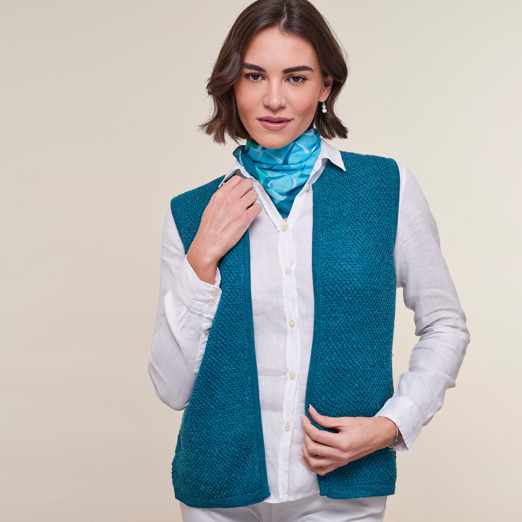A woman modeling a teal blue-green textured knit vest over a white button-up shirt, accessorized with a bright blue neck scarf, against a solid beige studio background.