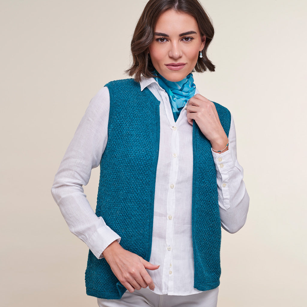 A woman modeling a teal textured knit vest over a white button-down shirt and a blue neck scarf, set against a solid light beige background.