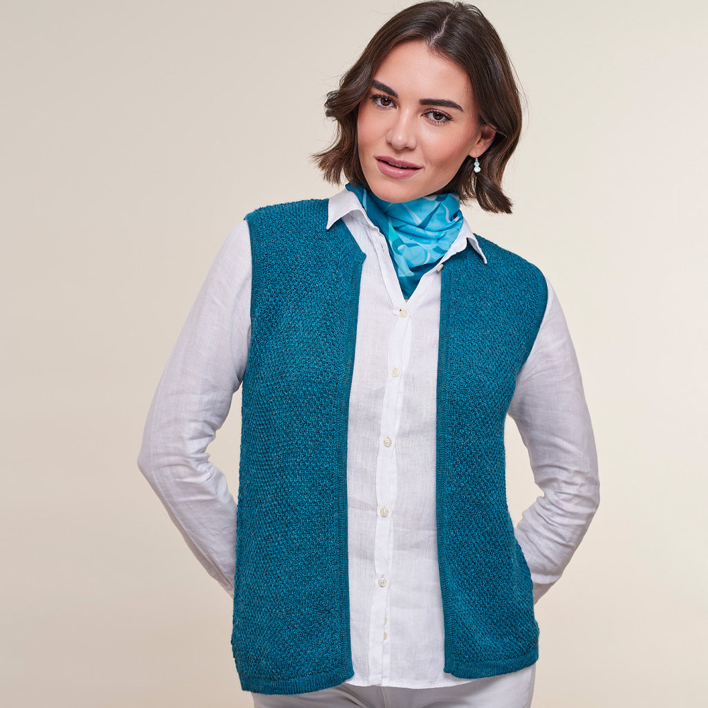 A woman modeling a teal blue textured knit vest over a white button-down shirt, accessorized with a bright blue neck scarf, against a solid light beige background.