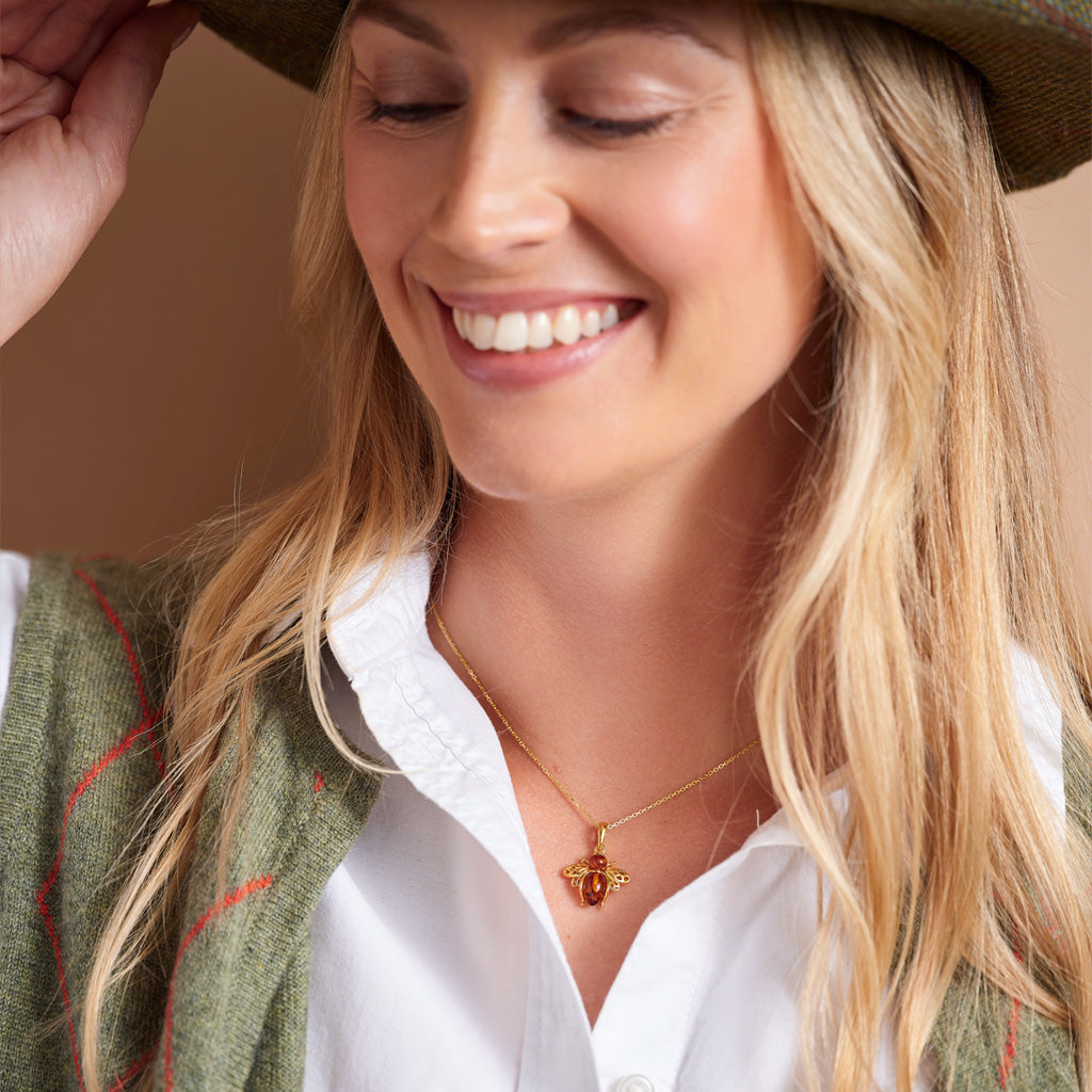 A smiling blonde woman wearing a delicate gold necklace with a small red bee pendant, styled with a green hat, white shirt, and green tweed cardigan.