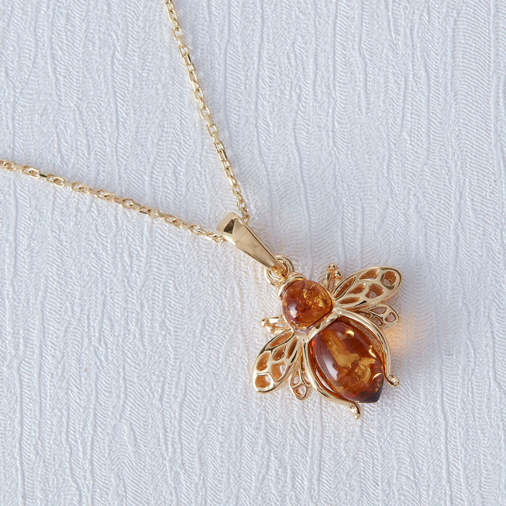 A gold-tone beehive pendant necklace featuring amber-colored gemstones, displayed on a textured white fabric background.