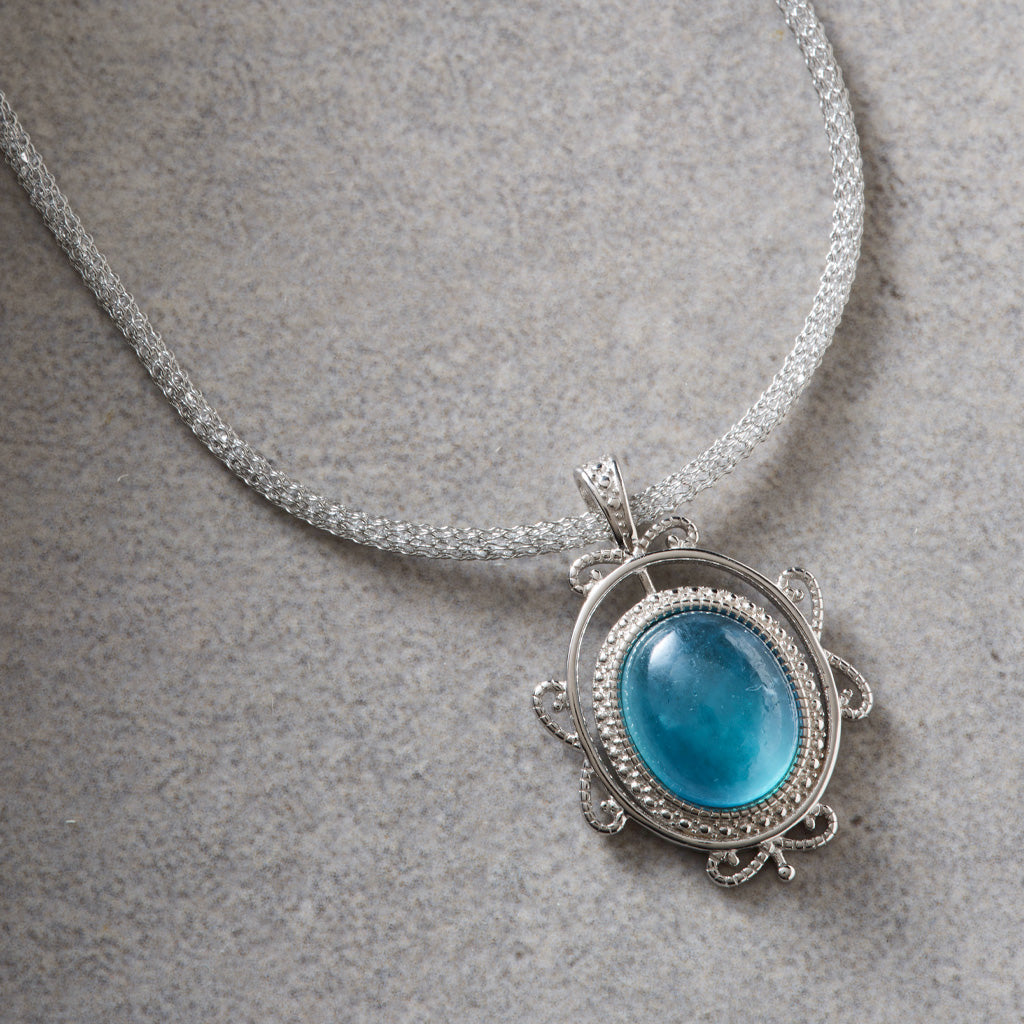 A silver-toned necklace with a textured chain and an oval pendant featuring a prominent light blue cabochon gemstone surrounded by a silver filigree border, displayed on a textured gray surface.