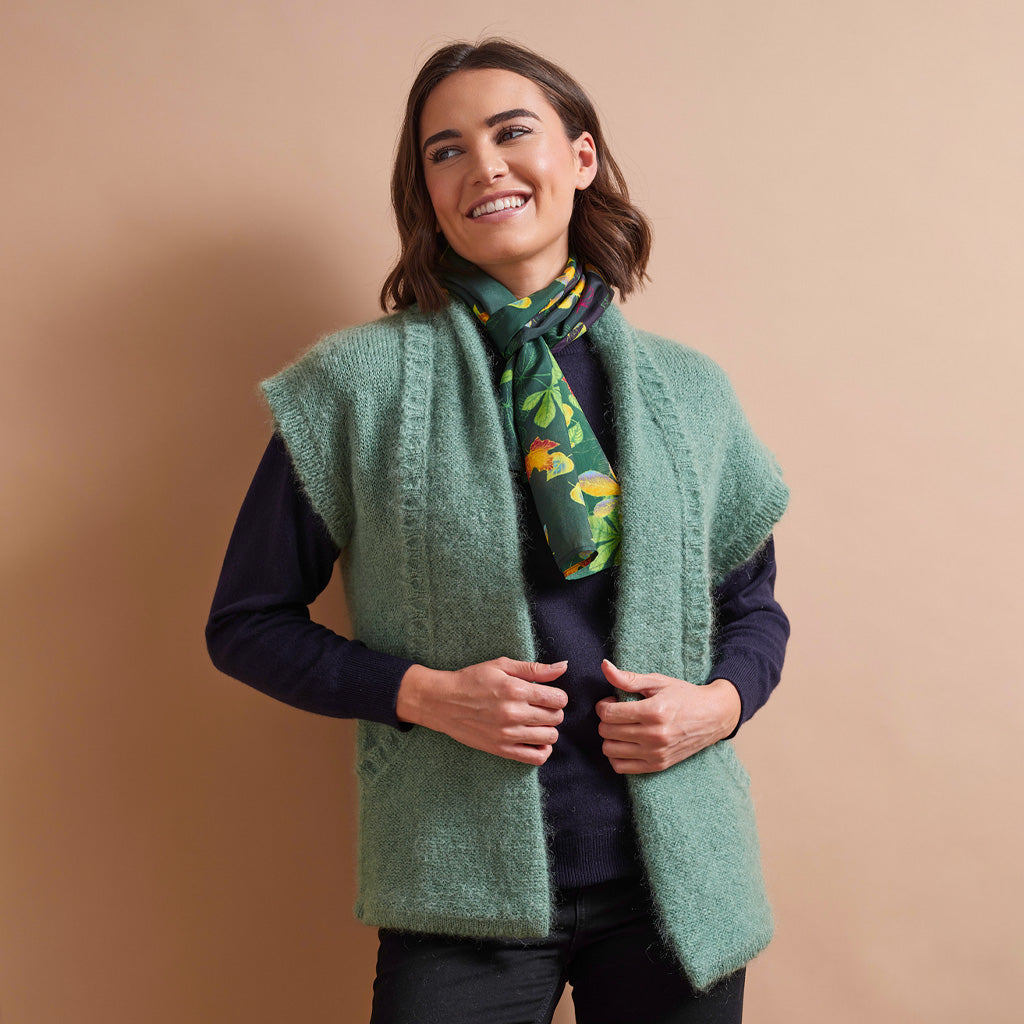 A woman wearing a fuzzy sage green sleeveless vest over a dark navy long-sleeve top, accessorized with a green floral scarf, posing against a solid beige background.
