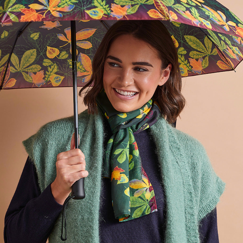 A woman smiling while holding an open umbrella, wearing a green knit sweater vest and a dark green scarf with a colorful autumn leaf pattern. The umbrella and scarf feature a matching leaf print on a dark background.