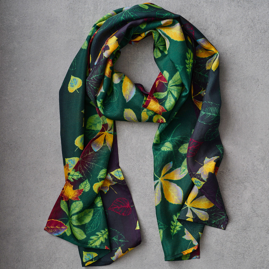 A dark green scarf with a vibrant multicolor botanical leaf print, draped in a loop on a textured gray background.