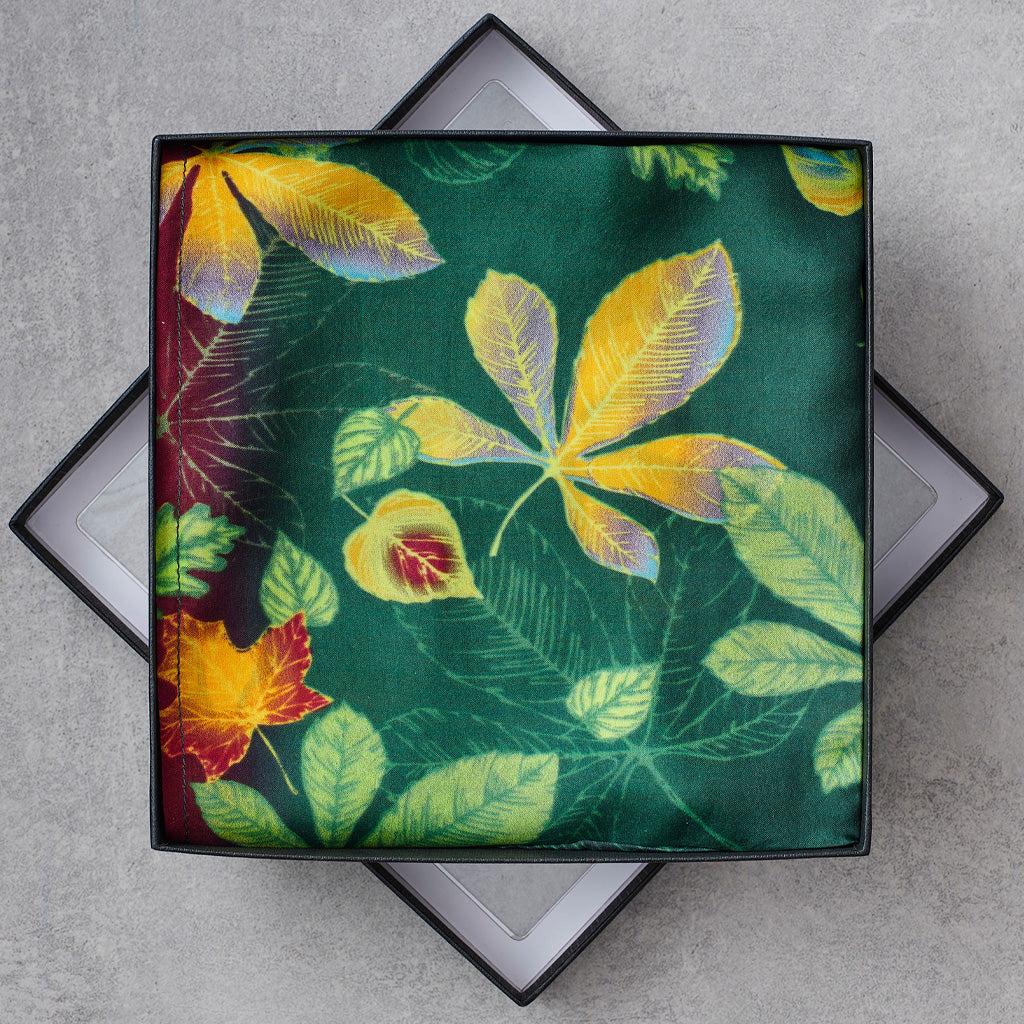 A square scarf with a botanical leaf pattern in green, yellow, and red, presented inside a black gift box on a gray textured background.