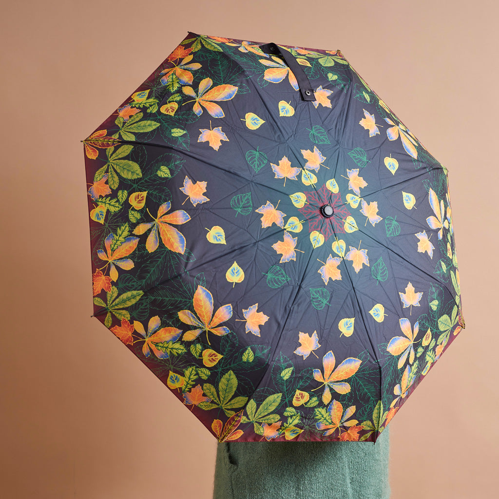An open umbrella with a dark navy background and a colorful print of autumn leaves in orange, yellow, green, and white, held up against a solid beige background.