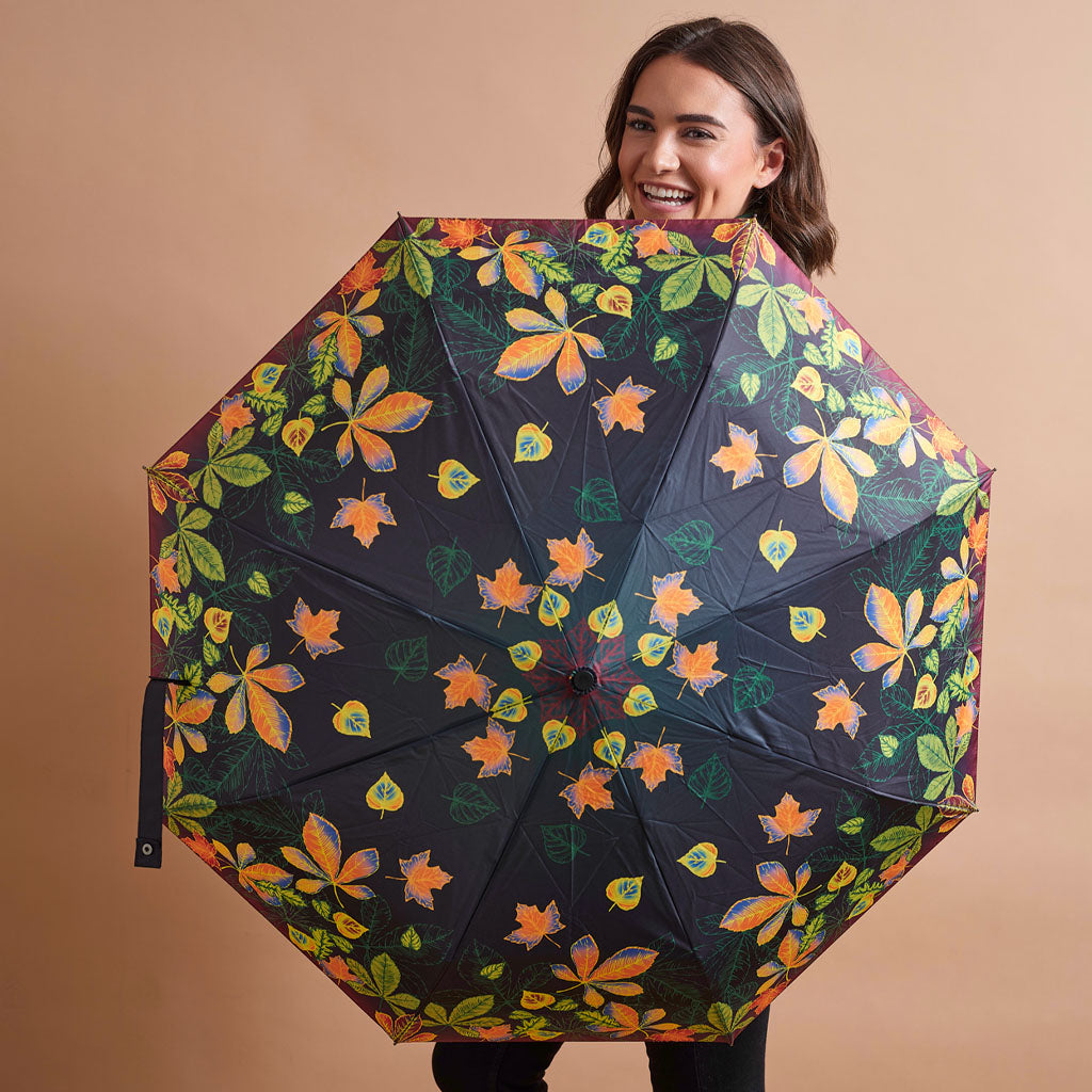 A woman holding up a large black umbrella with a vibrant autumn leaf pattern of orange, yellow, and green leaves, set against a solid beige background.