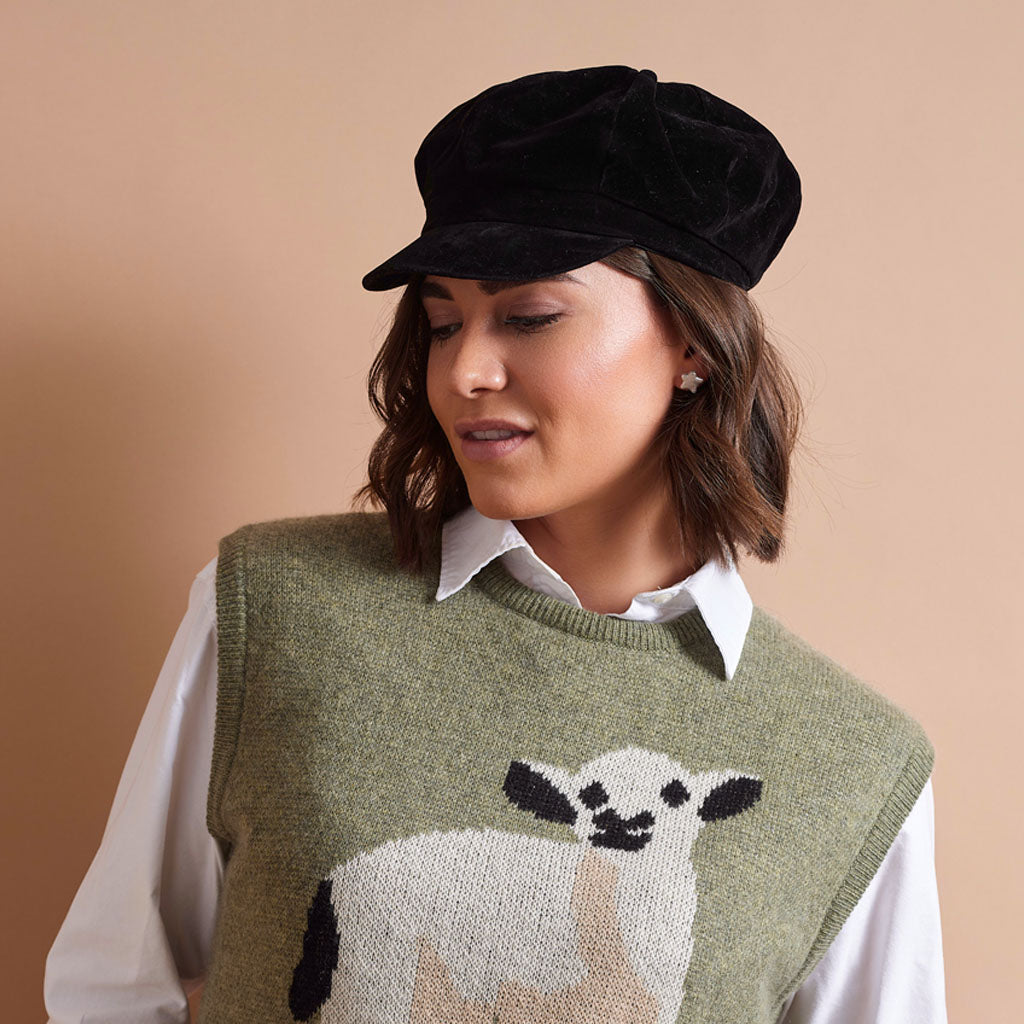 A woman wearing a black velvet newsboy cap and an olive green knit sweater vest with a white and black sheep motif, layered over a white collared shirt, against a solid beige background.