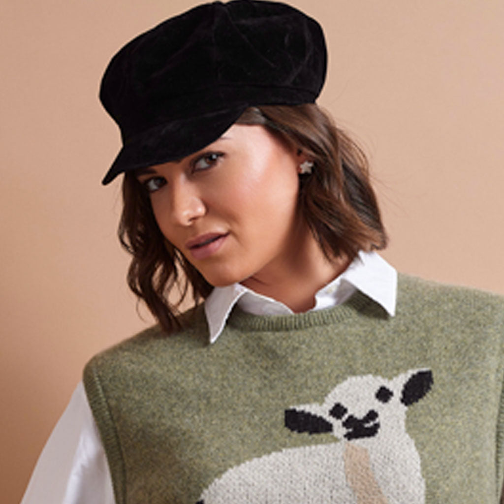 A woman wearing a black velvet newsboy cap, a white collared shirt, and an olive green knit sweater vest with a white and black sheep motif, set against a solid beige background.
