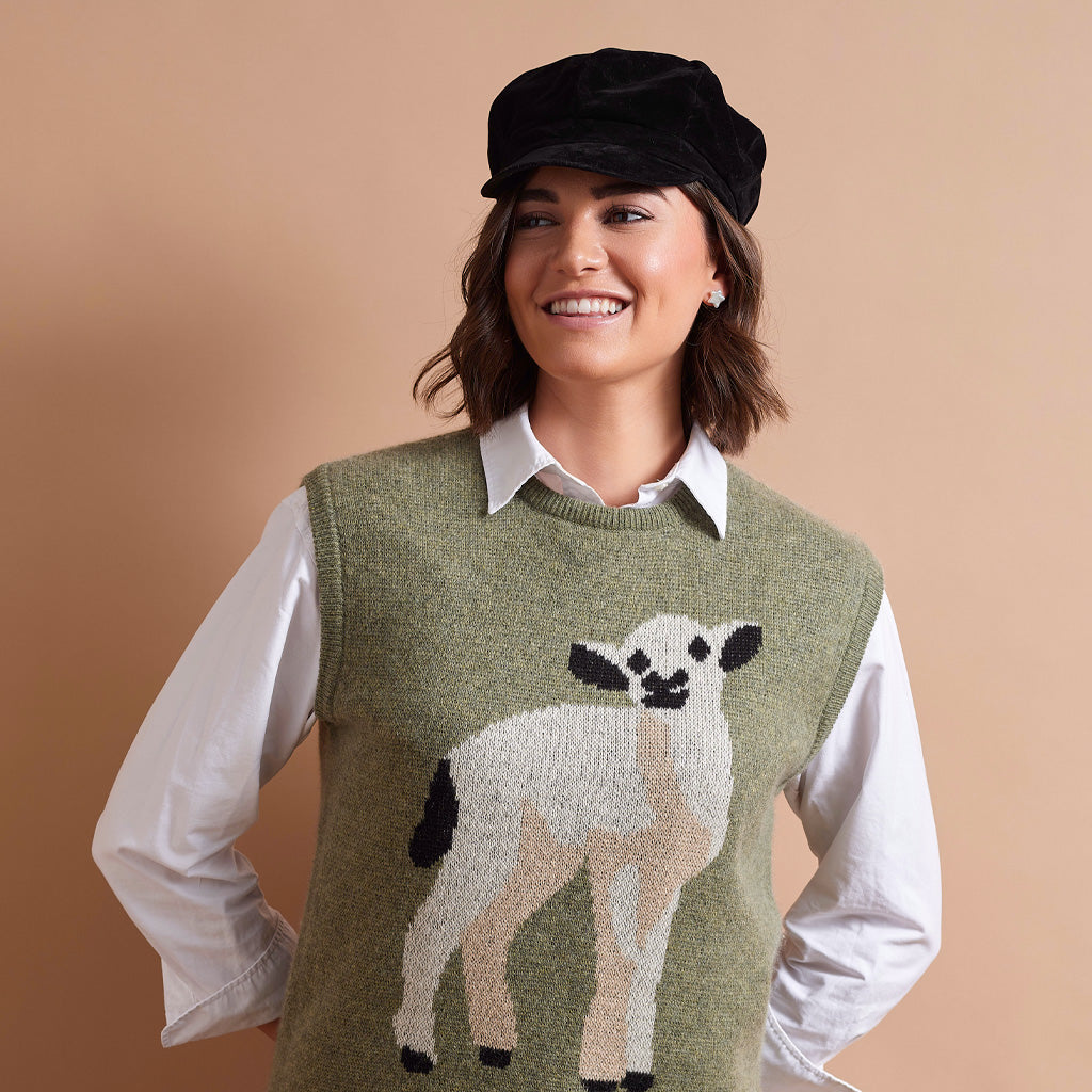 A woman wearing a black newsboy cap, a white collared shirt, and an olive green knit sweater vest with a large white, beige, and black lamb graphic on the front, posed against a solid warm beige background.