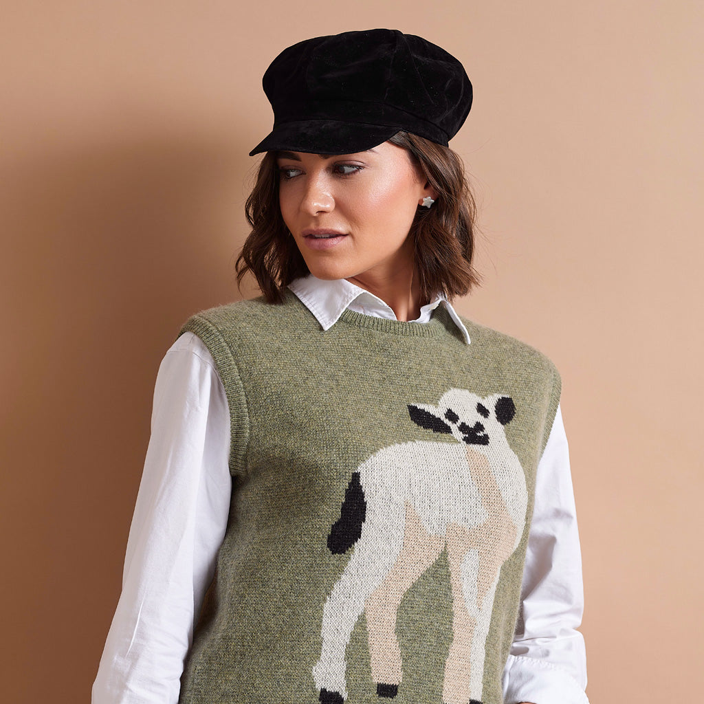 A woman wearing a black velvet newsboy cap, a white collared shirt, and an olive green knitted sweater vest with a large white, beige, and black lamb graphic on the front, set against a solid warm beige background.