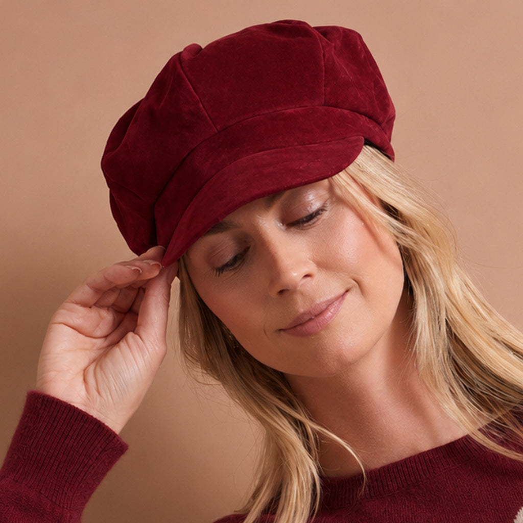 A woman wearing a deep burgundy velvet newsboy cap, adjusting the brim with her hand against a solid warm beige background.