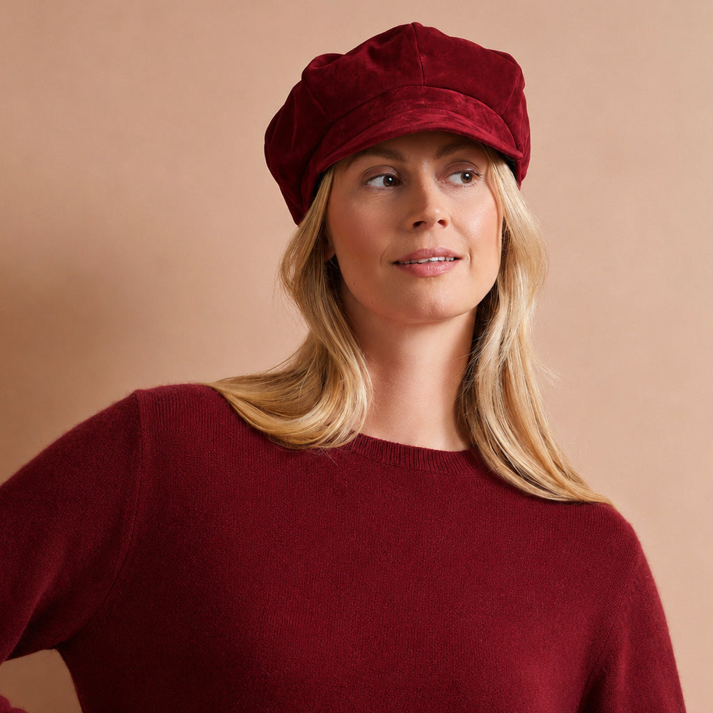 A woman wearing a deep burgundy velvet newsboy cap and a matching burgundy knit sweater, posed against a solid warm beige background.