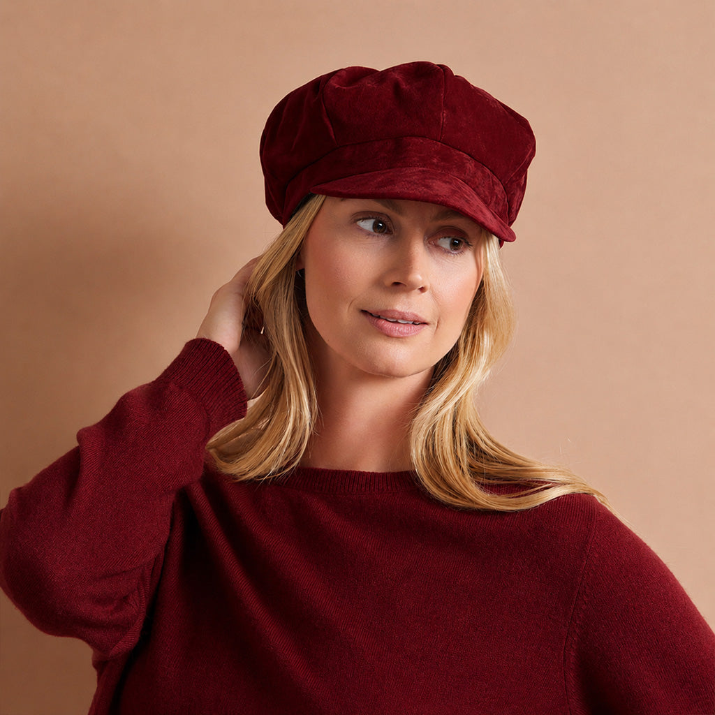 A woman wearing a deep burgundy velvet newsboy cap and a matching burgundy knit sweater, posed against a solid warm beige background.