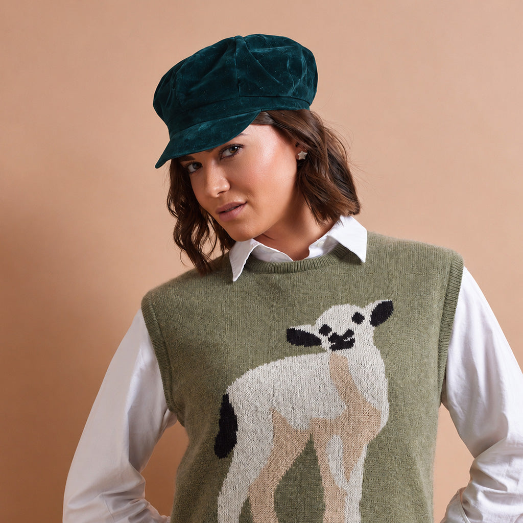 A woman wearing a dark green velvet newsboy cap, a white button-down shirt, and an olive green knit sweater vest with a knitted white and black lamb design on the front, posed against a solid beige background.