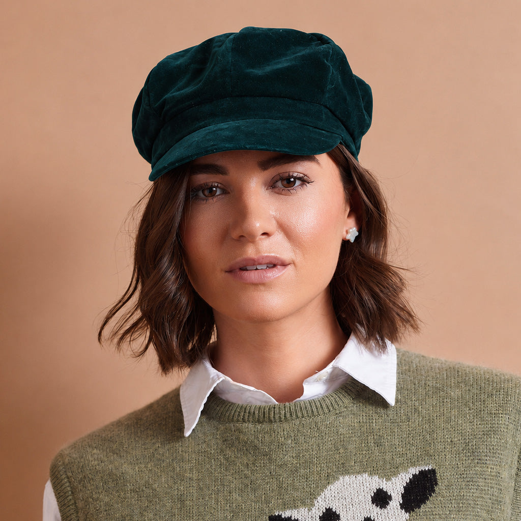 A woman wearing a dark green velvet newsboy cap and an olive green knit sweater over a white collared shirt, set against a solid warm beige background.