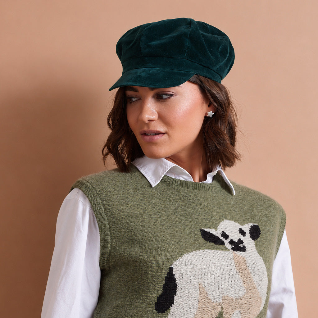 A woman wearing a dark green velvet newsboy cap, an olive green knit sweater vest with a sheep motif, and a white collared shirt, posed against a solid warm beige background.