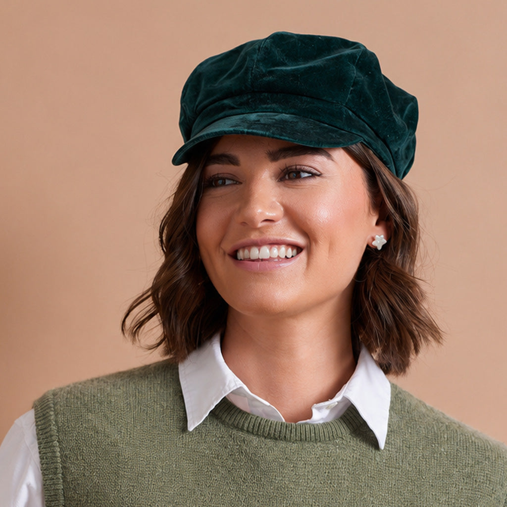 A woman wearing a dark green velvet newsboy cap, a white button-down shirt, and an olive green knit sweater vest with a knitted white and black lamb design on the front, posed against a solid beige background.