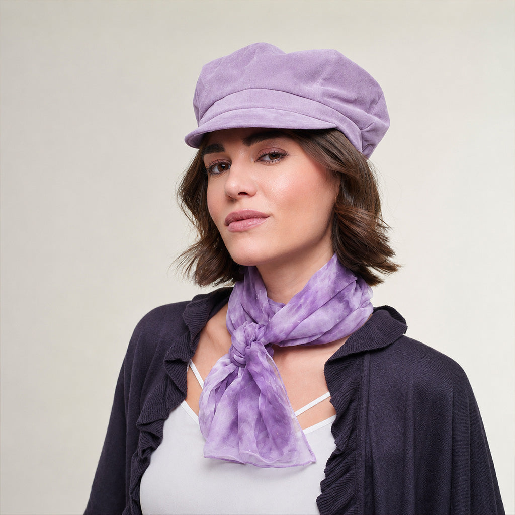 A woman wearing a lavender-purple newsboy cap and a matching purple tie-dye scarf, styled with a white top and a dark open-front cardigan against a plain light beige background.