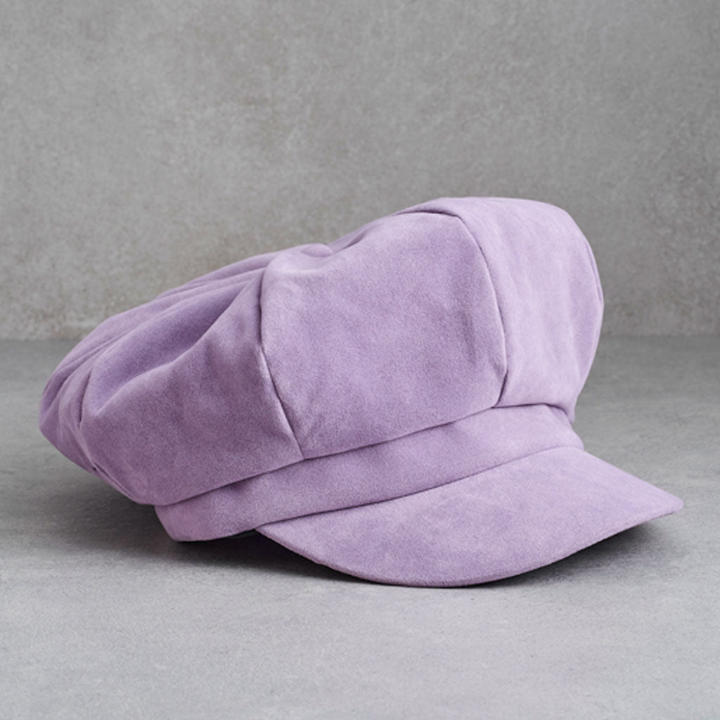 A lavender purple suede-like newsboy cap with a rounded crown and short brim, photographed on a neutral gray textured background.