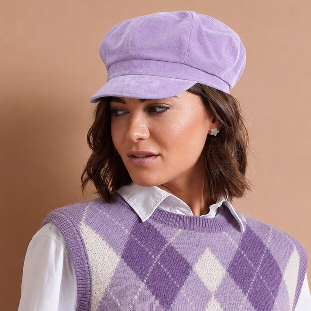 A woman wearing a lavender purple corduroy newsboy cap and a matching purple and white argyle knit sweater vest over a white collared shirt, set against a solid warm tan background.