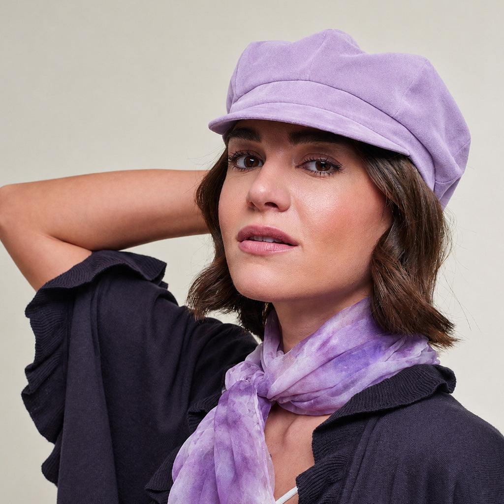 A woman wearing a lavender purple newsboy cap and a matching purple tie-dye scarf, paired with a dark navy cardigan, set against a plain light beige background.