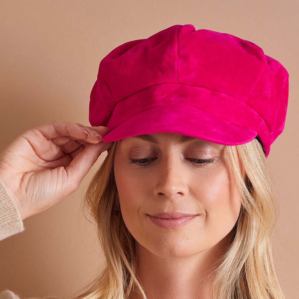 A woman wearing a bright hot pink velvet newsboy cap, adjusting the brim with her hand against a solid beige background.