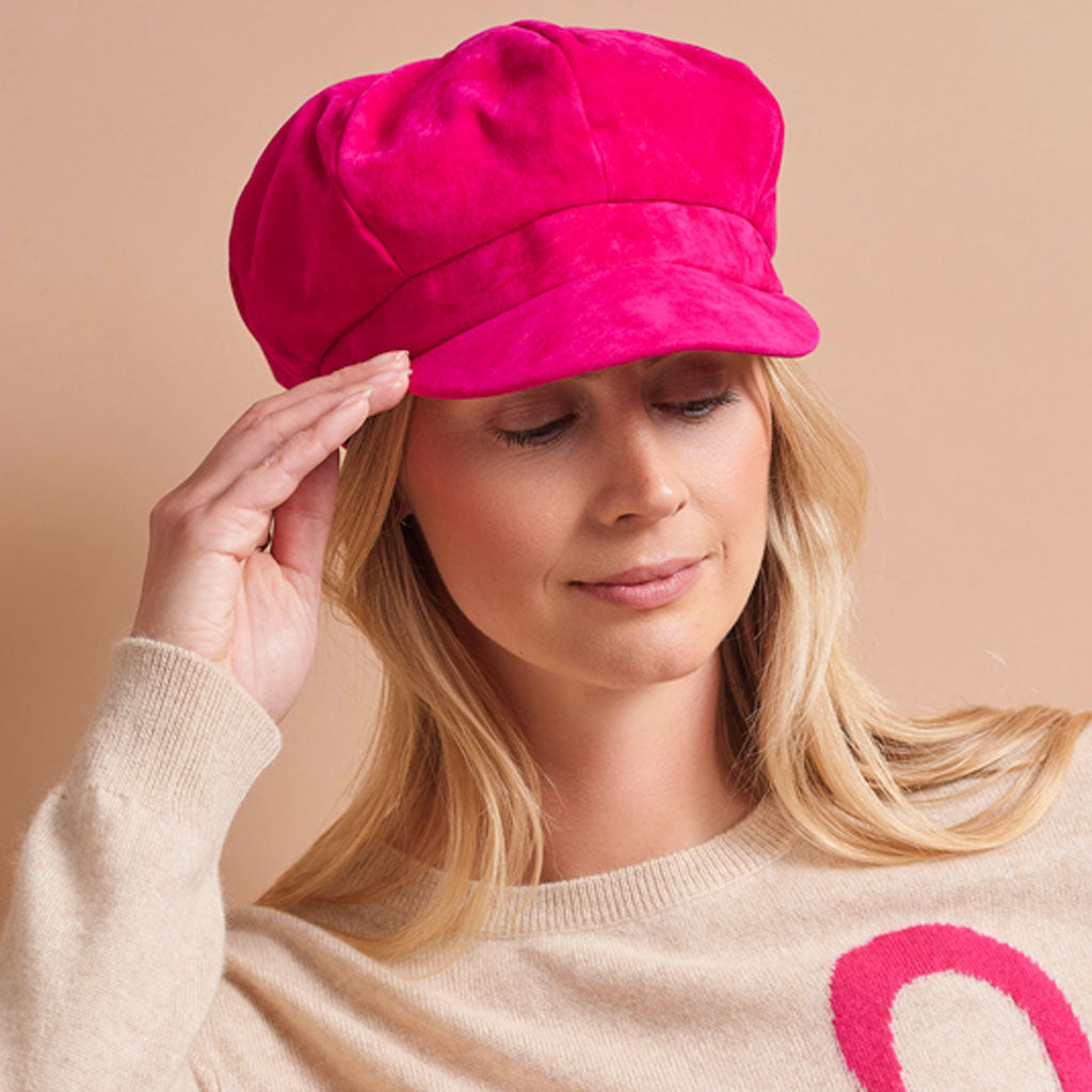 A woman wearing a bright hot pink velvet newsboy cap, touching the brim with her hand, against a solid beige background.