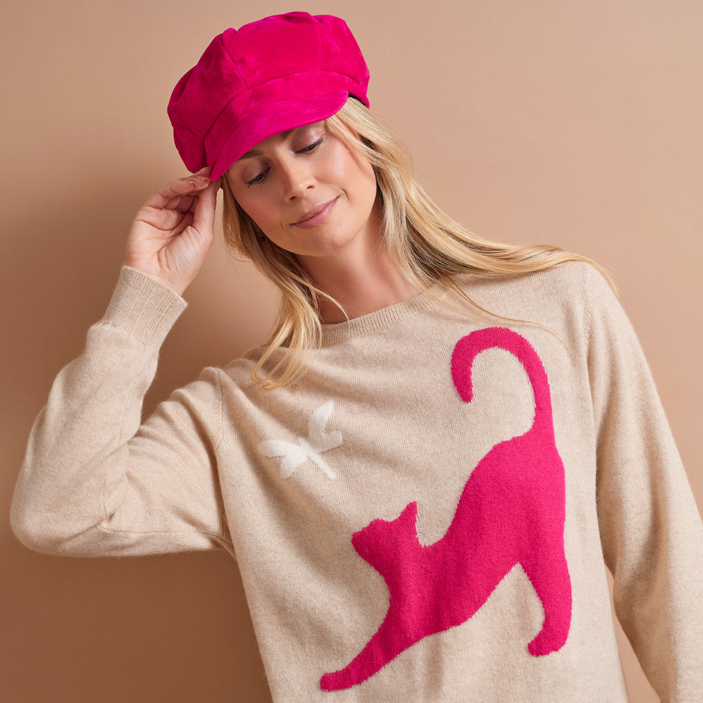 A woman wearing a bright pink newsboy cap and a beige knit sweater with a large pink cat silhouette and a small white bow on the chest, posed against a solid beige background.