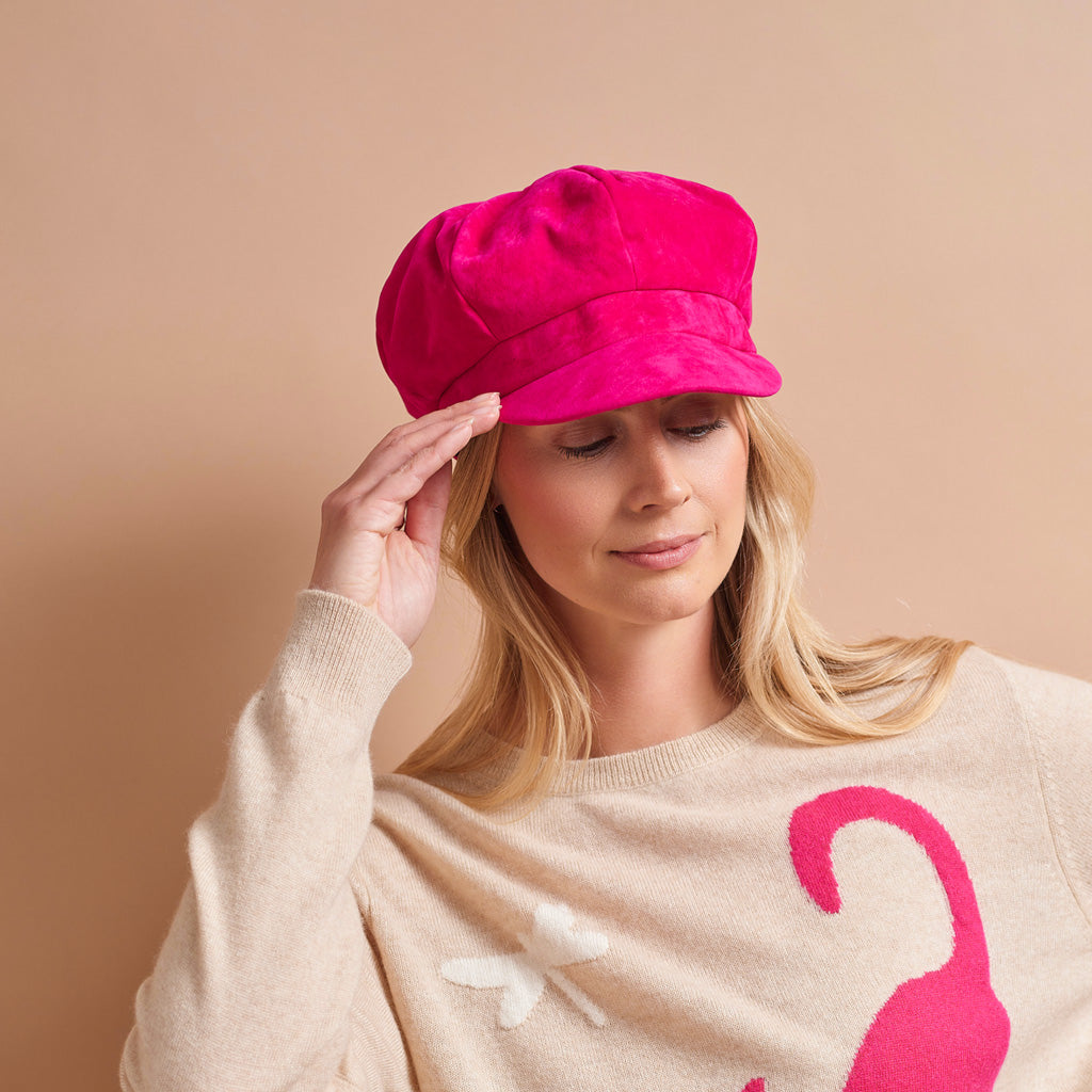 A woman wearing a bright pink velvet newsboy cap and a beige sweater with a pink flamingo graphic, posing against a solid beige background.