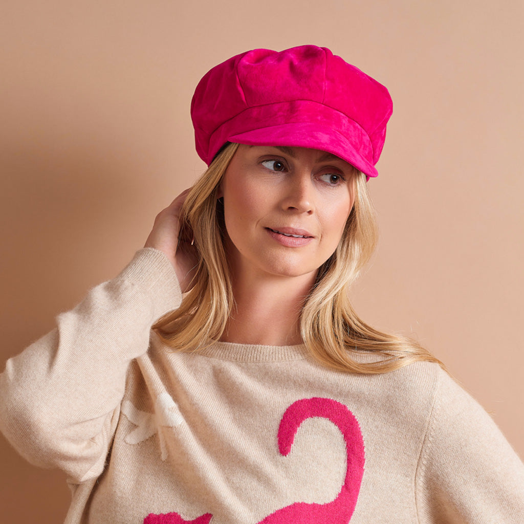A woman wearing a bright pink newsboy cap and a beige sweater with a pink graphic, posed against a solid beige background.