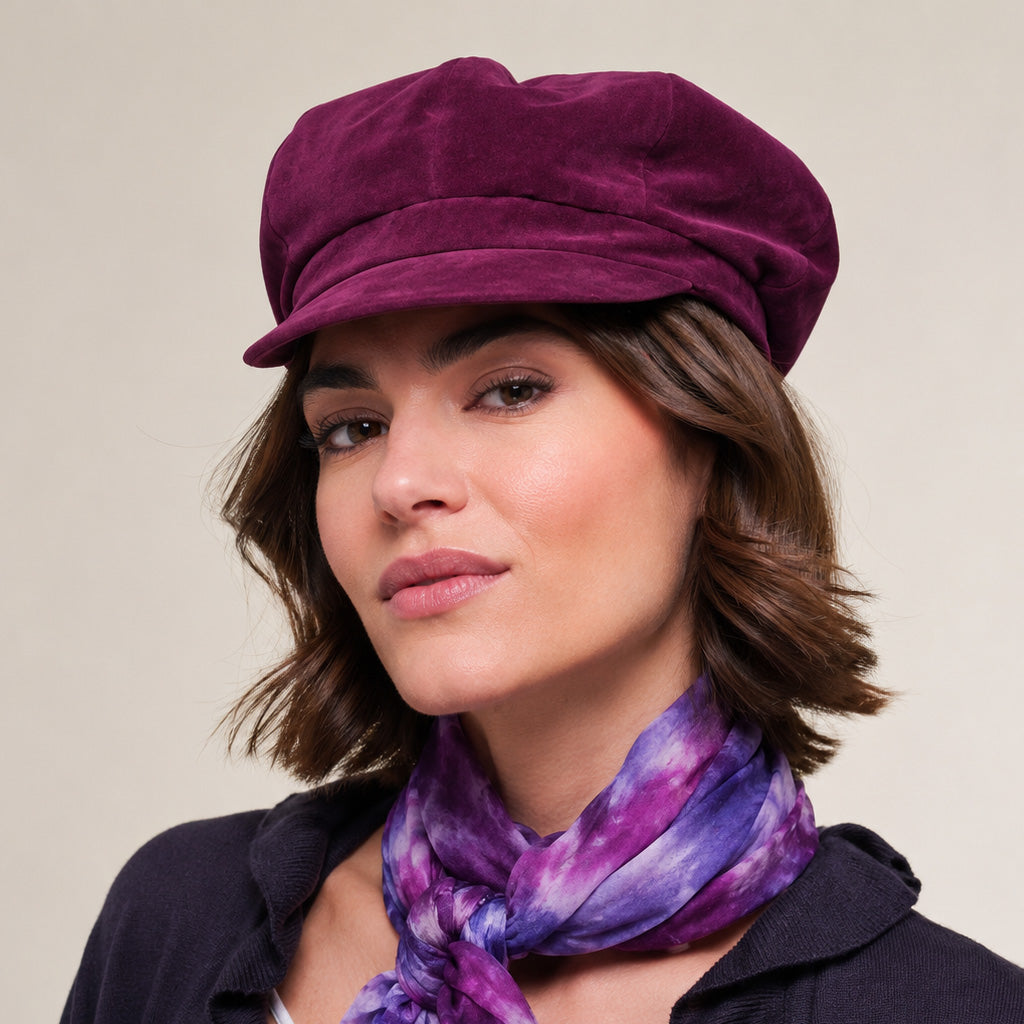 A woman wearing a deep purple velvet newsboy cap and a matching purple and blue tie-dye scarf, photographed against a solid light beige background.